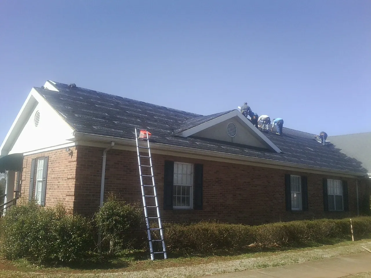 Crew tearing shingles off a brick ranch roof with a ladder set — Emergency Roof Repair work by our crew in Laurel Hill